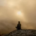 Image of a person sat at the top of a hill where the sun is barely visible through the clouds to illustrate post